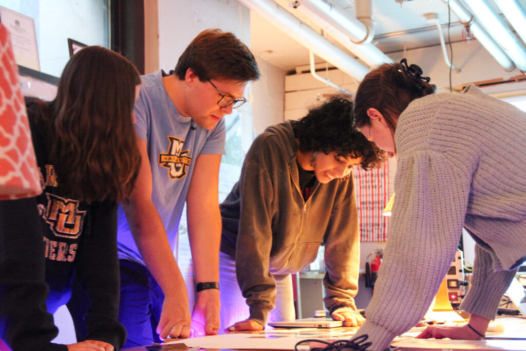 four students lean over a table in their workspace on campus to discuss a recent project.