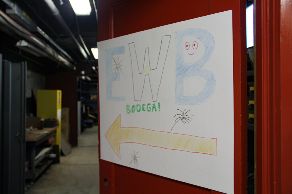 A slightly open door with a hand-written poster taped on it that reads "EWB Bodega!" with an arrow inviting people down a hallway.