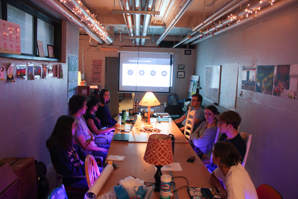 Students collaborate around a long meeting table in a simple room with club decor and keepsakes on the wall.