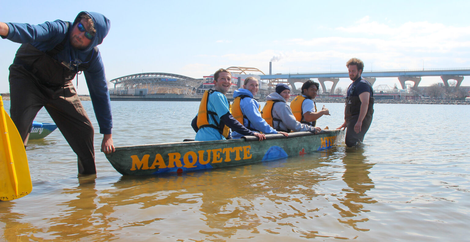 Four students in a concrete canoe, guided by two students in the water, with Milwaukee's Hoan Bridge and Summerfest grounds in the background