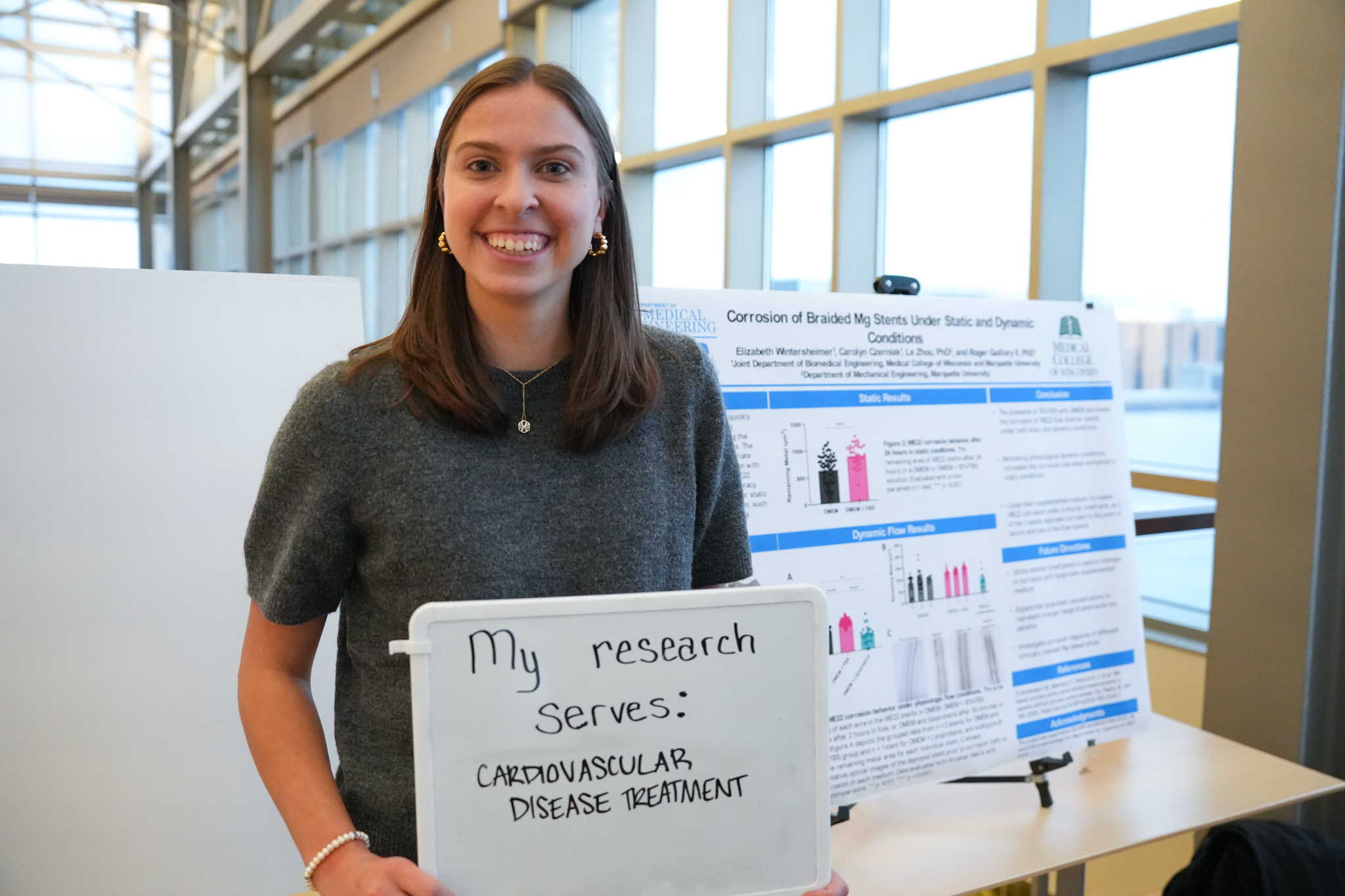 student holding a board that says "my research serves: cardiovascular disease treatment" in front of a research poster