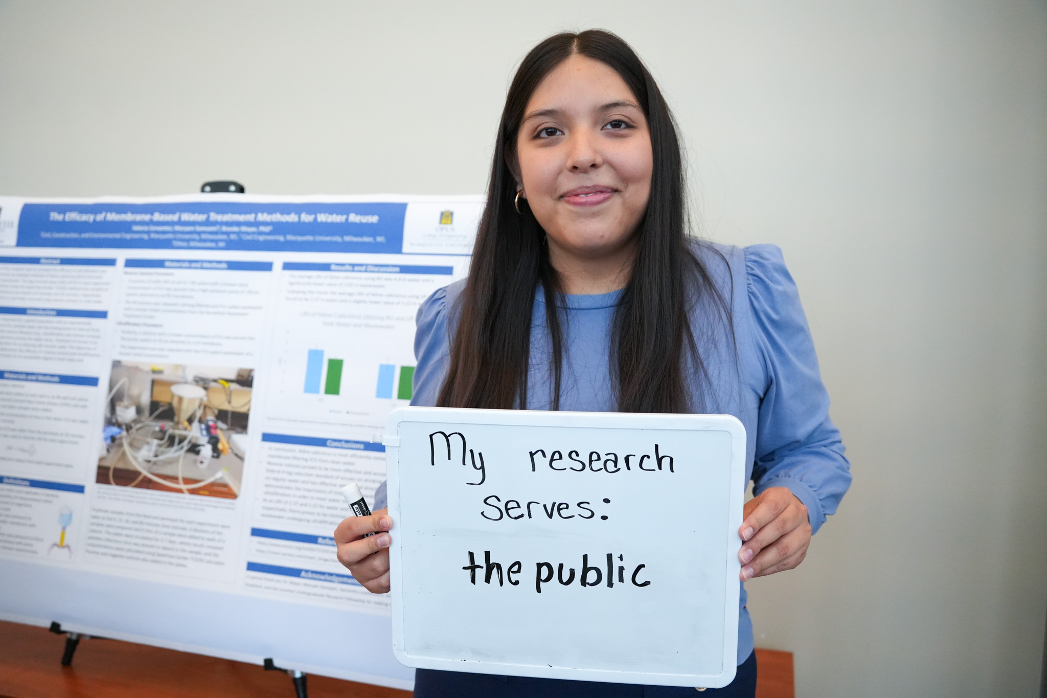 student holding a board that says "my research serves: the public" in front of a research poster
