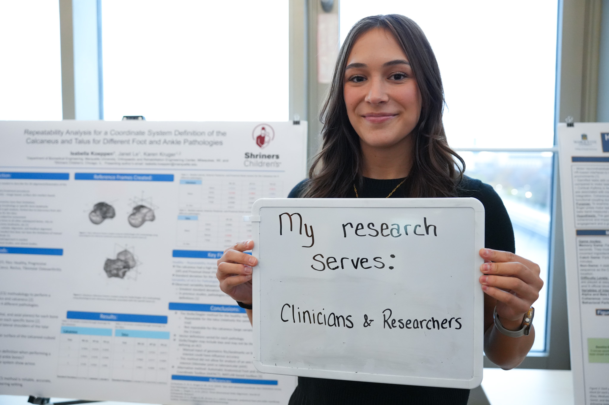 student holding a board that says "my research serves: clinicians & researchers" in front of a research poster
