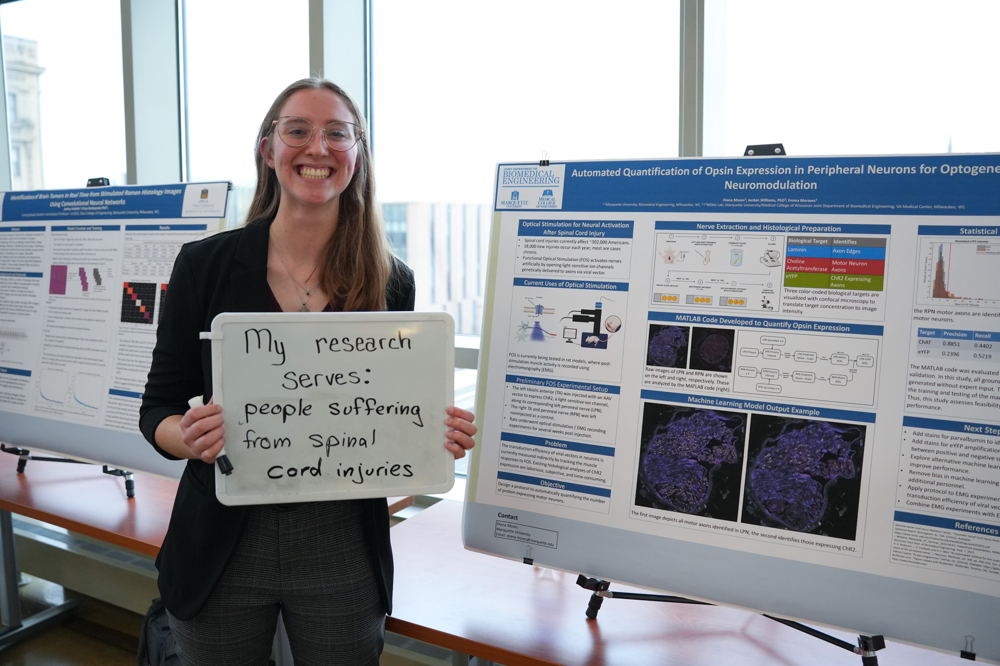 student holding a board that says "my research serves: people suffering from spinal cord injuries" in front of a research poster