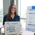 student holding a board that says "my research serves: people with spinal cord injuries" in front of a research poster