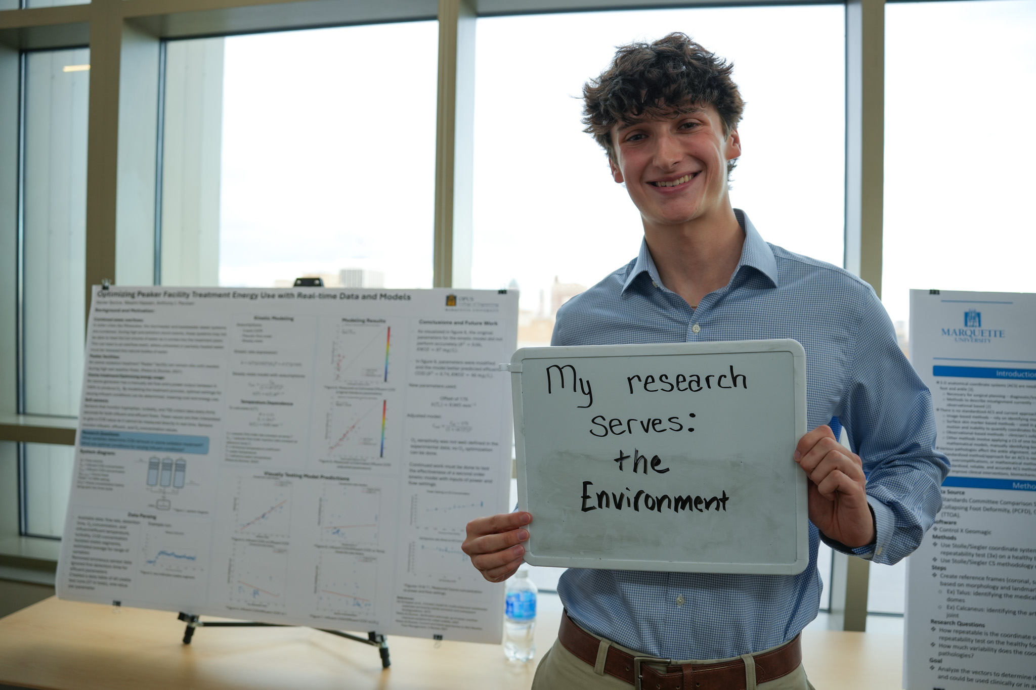student holding a board that says "my research serves: the environment" in front of a research poster