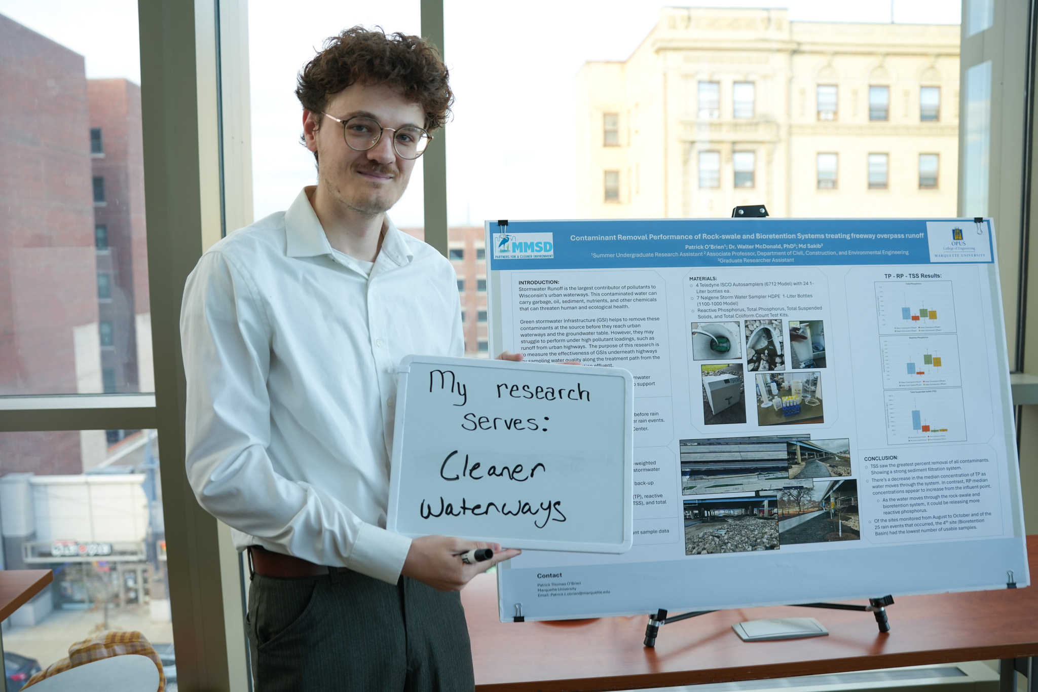 student holding a board that says "my research serves: cleaner waterways" in front of a research poster