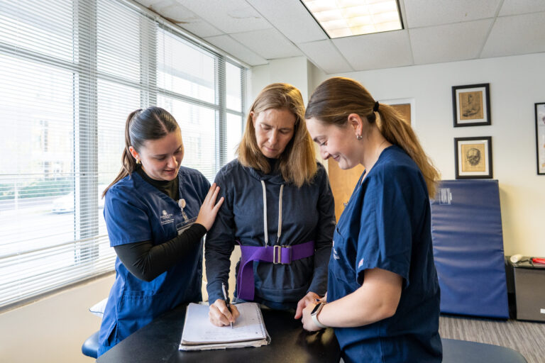 Two students assisting a patient write on a notepad.