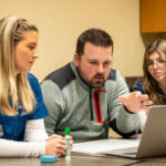 A man speaks while reading a prompt off a computer at the direction of students.