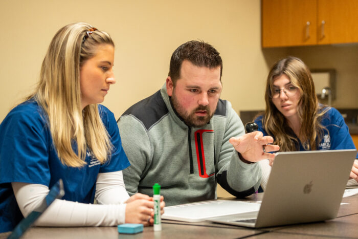 A man speaks while reading a prompt off a computer at the direction of students.