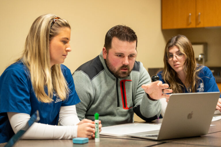A man speaks while reading a prompt off a computer at the direction of students.