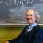 Dr. Richie sitting at desk with chalkboard behind him
