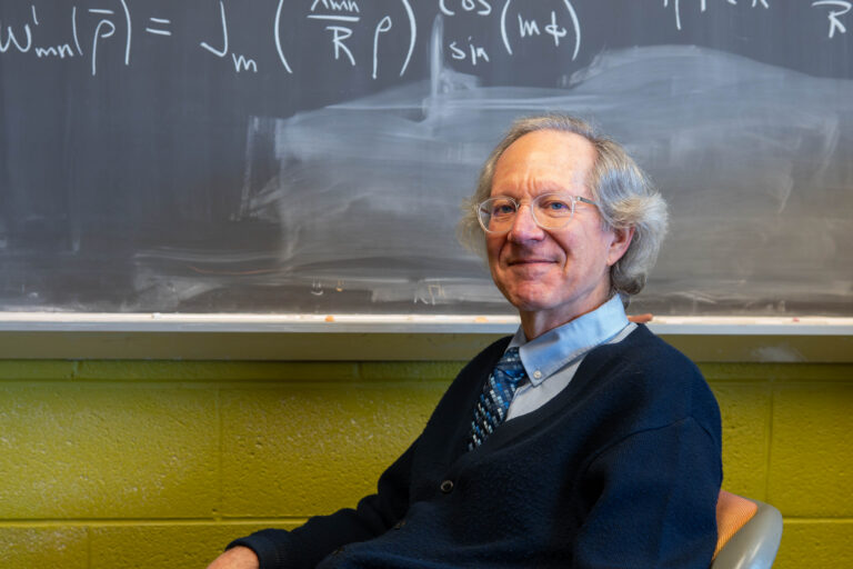 Dr. Richie sitting at desk with chalkboard behind him