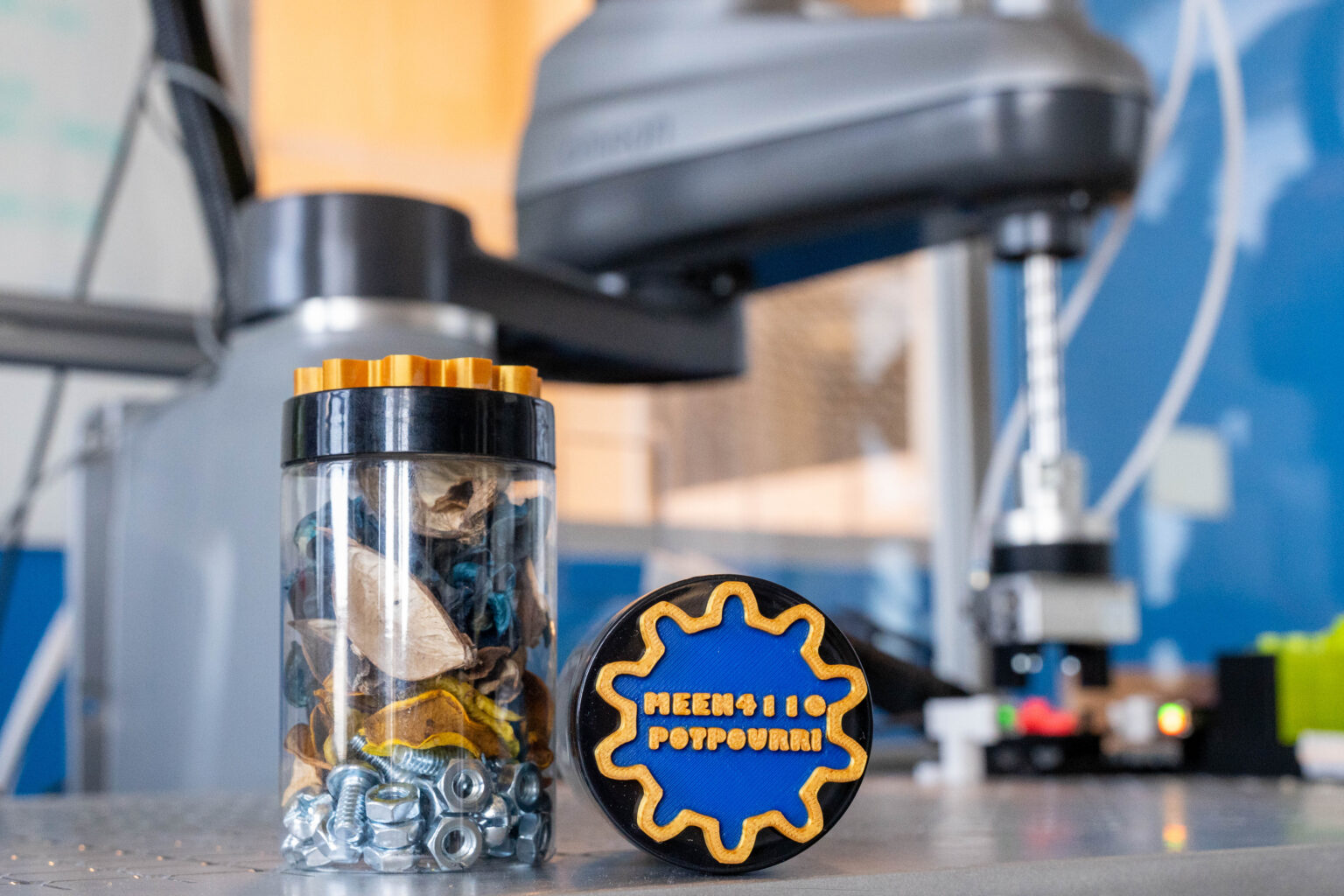 Blue and gold potpourri containers labeled "MEEN4110 Potpourri" in front of a robotic arm lab experiment