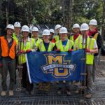 students wearing hard hats and high visibility vests posing with a Marquette flag on the foundation of a water tank construction