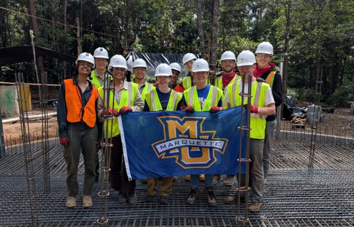 students wearing hard hats and high visibility vests posing with a Marquette flag on the foundation of a water tank construction